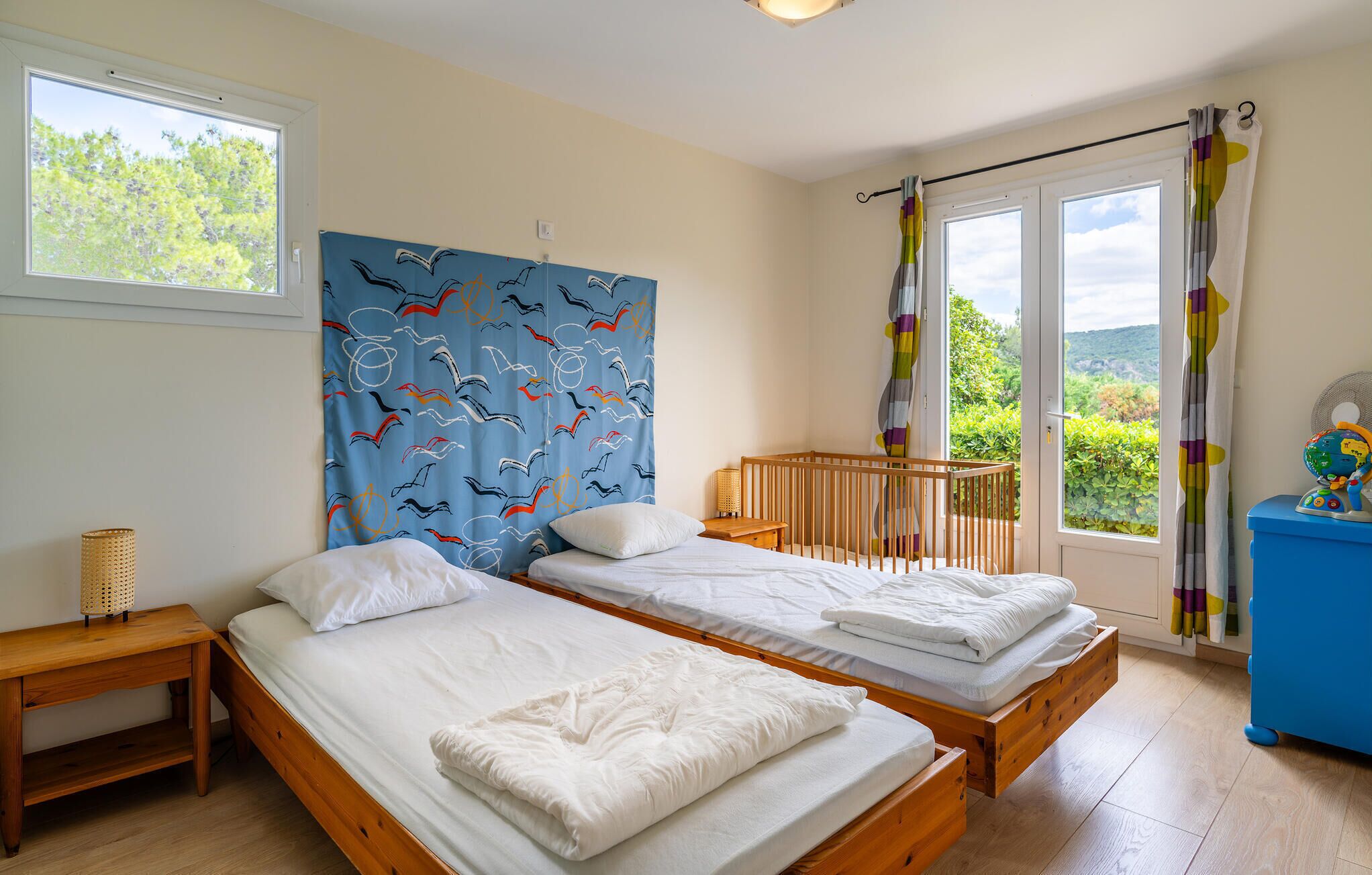 A bedroom with two beds, a crib, a window with a view of greenery, and a blue curtain with a bird pattern.