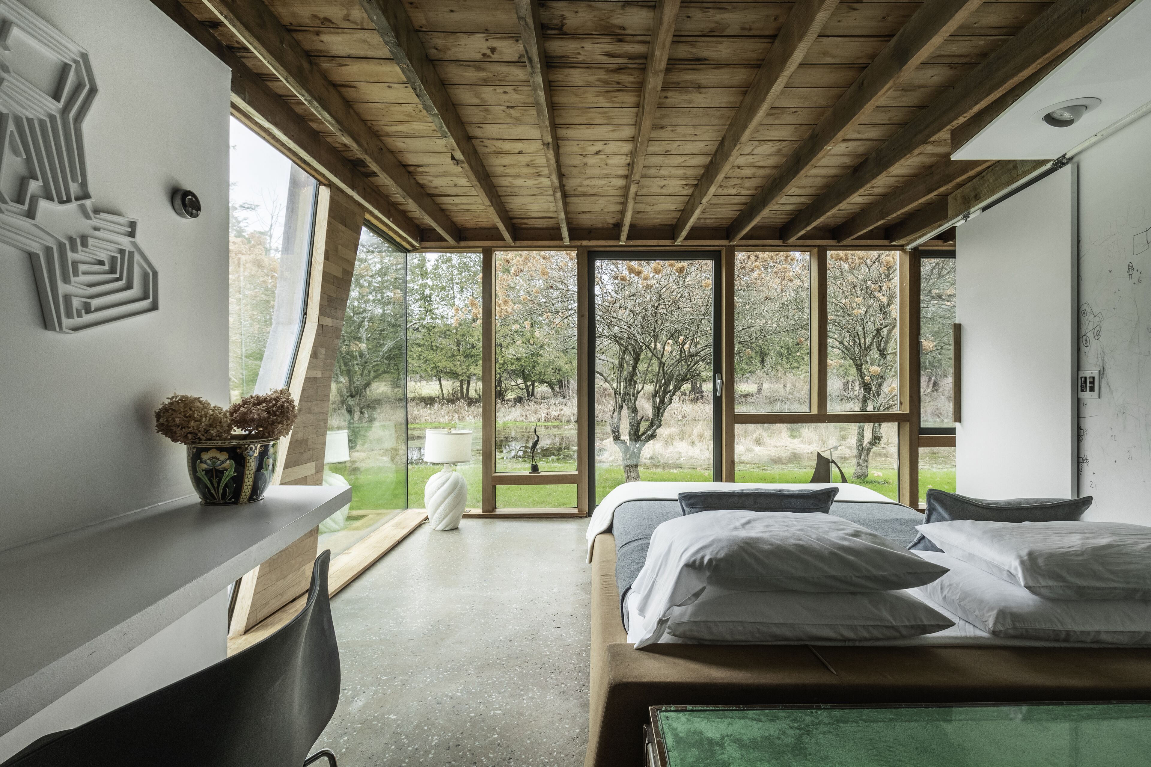 A modern bedroom with a wooden ceiling, large windows, a bed with white linens, and a small table with a vase.