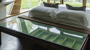 Un dormitorio con cama de cristal, ventanas grandes y vistas a árboles.