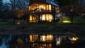 Una casa moderna con ventanas amplias, una chimenea de piedra y un reflejo en un estanque.