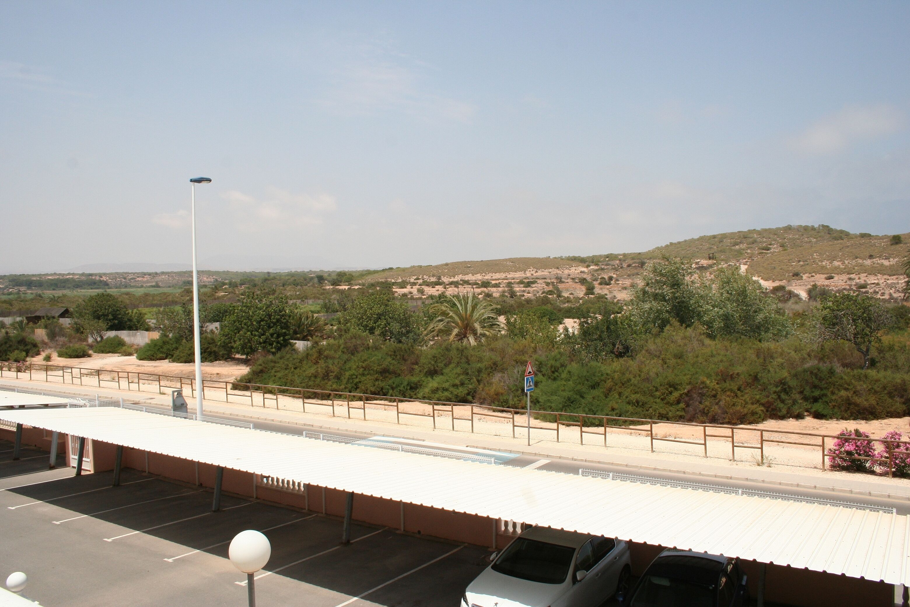 A parking area with a covered waiting area, a streetlight, and a view of a hilly landscape with sparse vegetation.