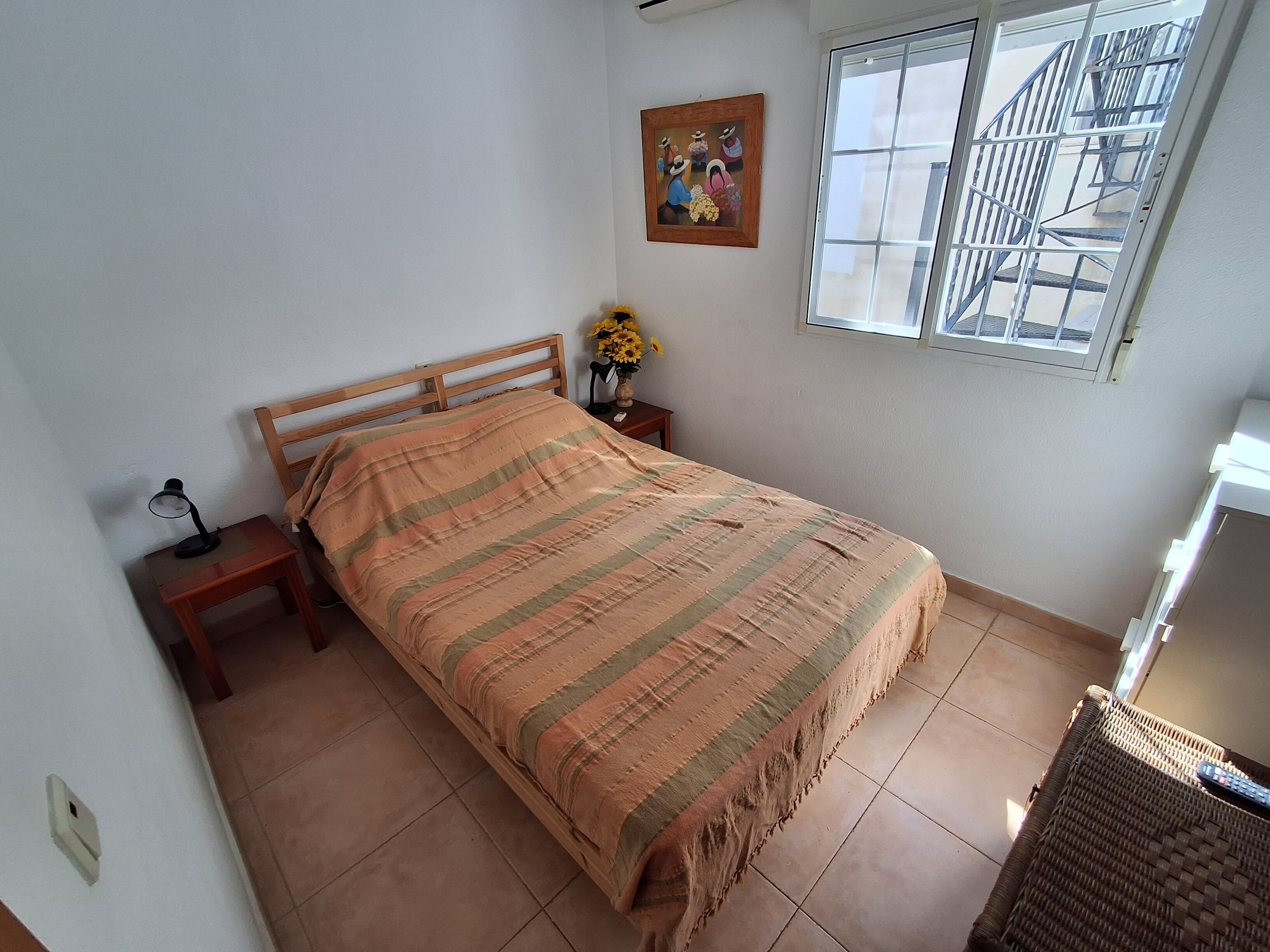 Un dormitorio con cama, mesita de noche, lámpara, un cuadro y una ventana.