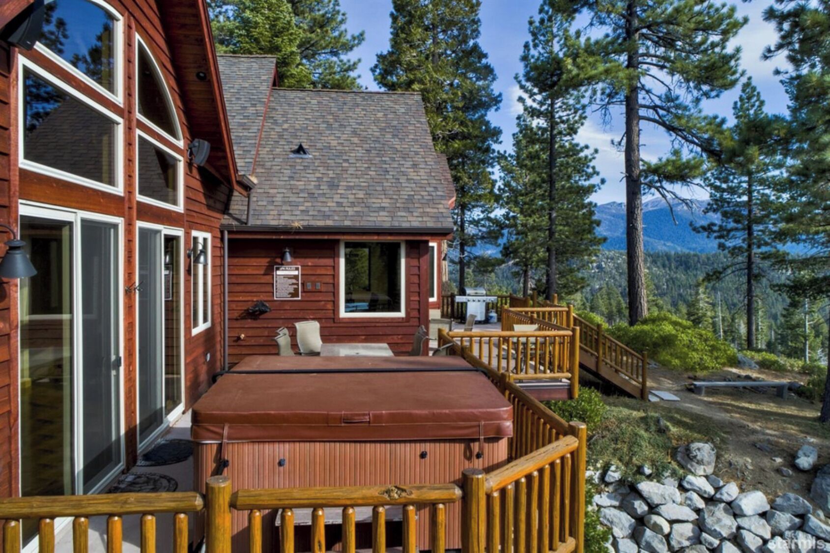 A wooden cabin with a hot tub on a deck, surrounded by trees and mountains.