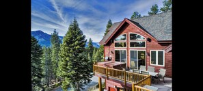 A wooden cabin with a deck, large windows, and a mountain backdrop.