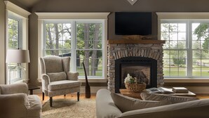 A living room with a stone fireplace, large windows, a comfortable armchair, and a sofa.