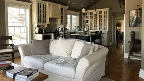 Un salon spacieux avec un canapé blanc, une table basse sur laquelle sont posés des livres, et une cuisine en arrière-plan, avec des meubles blancs et une grande fenêtre.