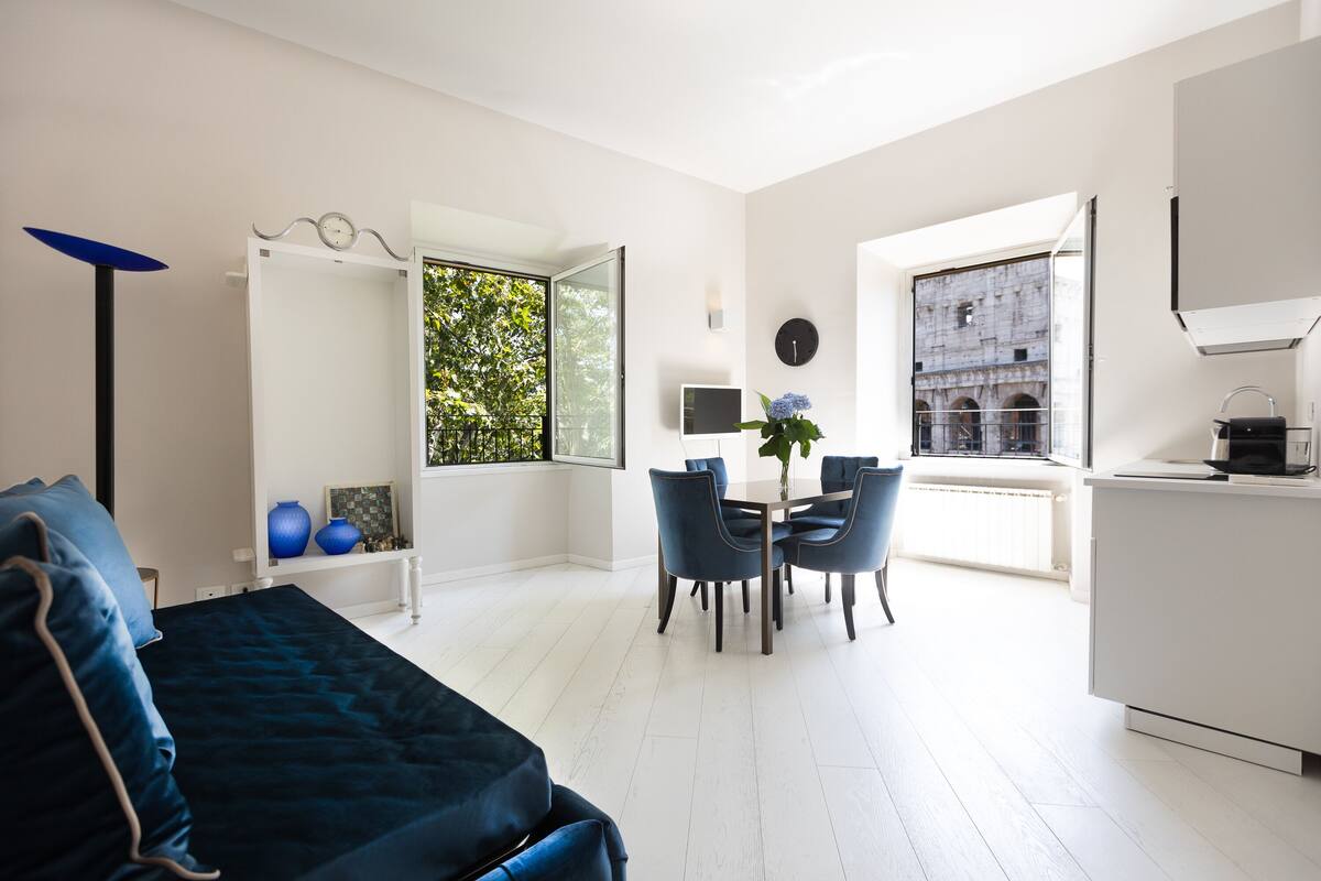 A modern, well-lit room with a bed, a dining table with chairs, a small kitchen area, and a large window with a view of greenery.