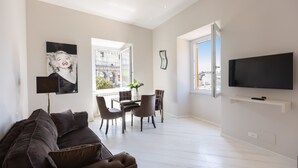 A modern living room with a dark brown sofa, a dining table with chairs, a flat-screen TV mounted on the wall, and a large window offering a view of the cityscape. - Amazing Colosseo (Rome)