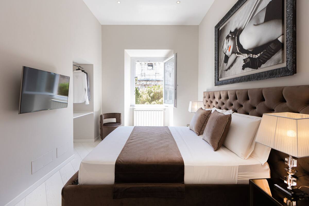 A modern hotel room with a large bed, a flat-screen TV, a framed artwork, and a view of the outdoors.