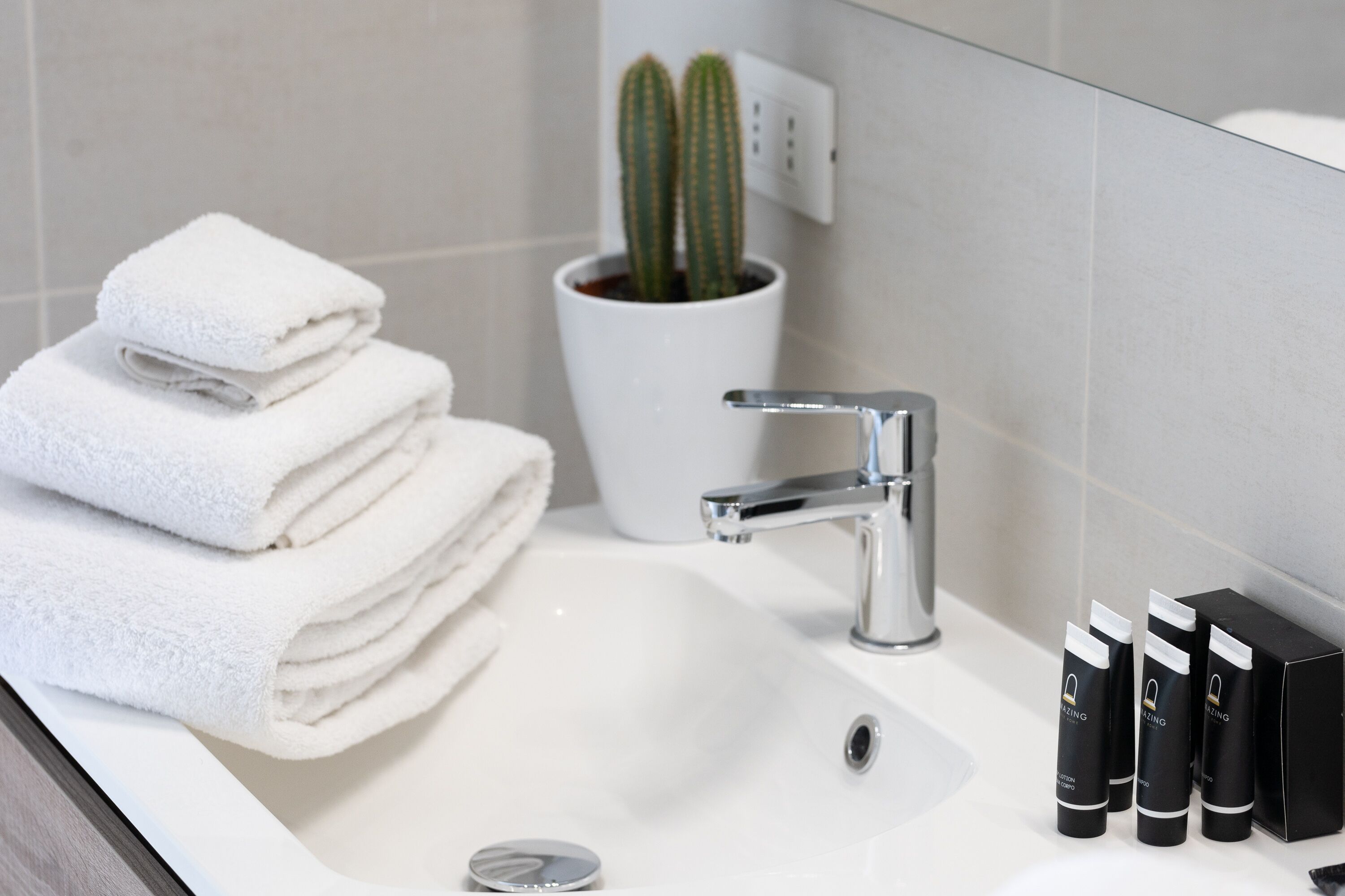 Un bagno con un lavandino bianco, uno specchio, un rubinetto, un mucchio di asciugamani bianchi, un cactus in vaso e una fila di articoli da toilette neri.