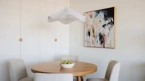 Ein Essbereich mit einem runden Holztisch, zwei Stühlen, einer weißen Schüssel mit Birnen, einer hängenden weißen Leuchte und einem großen abstrakten Gemälde an der Wand.