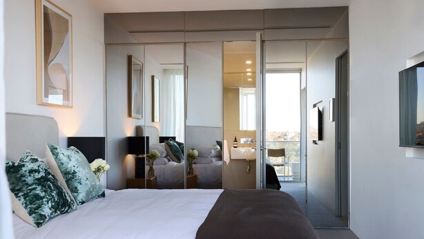 A modern hotel room with a large bed, a walk-in closet, a bathroom with a sink and mirror, and a view of the city through the window. - Sky Pad by Contemporary Hotels (DARLINGHURST)