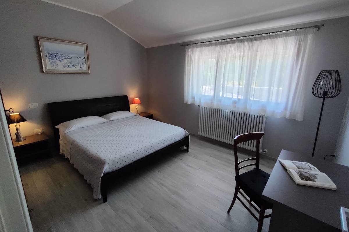 A bedroom with a bed, bedside lamps, a chair, a desk, a radiator, and a window with curtains.