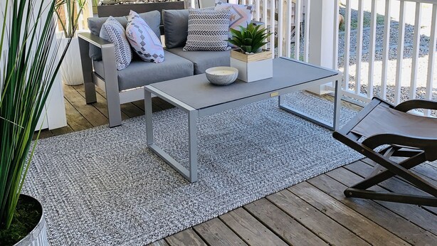 Een overdekt terras met een grijze bank, een witte salontafel en een kamerkleed met dessin.