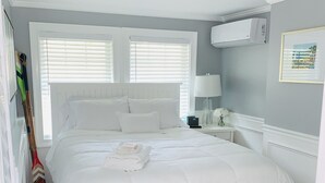 Een net opgemaakt bed met wit beddengoed, een nachtkastje met een lamp en een wandgemonteerde airconditioner.