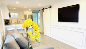 Een moderne woonkamer met een grijze bank, een witte bijzettafel met een gele orchidee en een flatscreen televisie aan de muur.