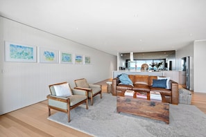 Un salon moderne avec un canapé, des fauteuils et une table basse. On aperçoit une cuisine au fond, avec vue sur la mer.
