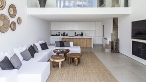 A modern living room with a white sofa, wooden coffee tables, and a kitchen in the background. - The Margi Villa (Vari-Voula-Vouliagmeni)