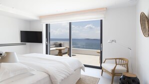 A modern bedroom with a large bed, a flat-screen TV, a wooden chair, and a view of the sea through large glass doors. - The Margi Villa (Vari-Voula-Vouliagmeni)