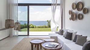 A modern living room with a white sofa, two wooden side tables, and a view of the sea through large glass doors. - The Margi Villa (Vari-Voula-Vouliagmeni)