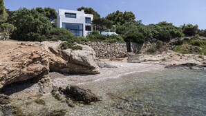 A modern house by a rocky beach with clear water, surrounded by trees. - The Margi Villa (Vari-Voula-Vouliagmeni)