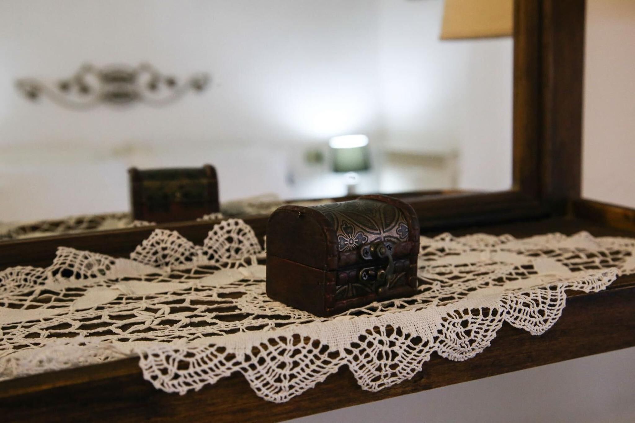 A small, ornate chest placed on a lace doily, with a mirror reflecting a lamp and a framed picture in the background.