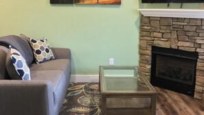 A living room with a sofa, a glass-top coffee table, a fireplace, and a mounted TV. - Blue Ridge Cottage Walking Distance to Downtown Waynesville (Waynesville)