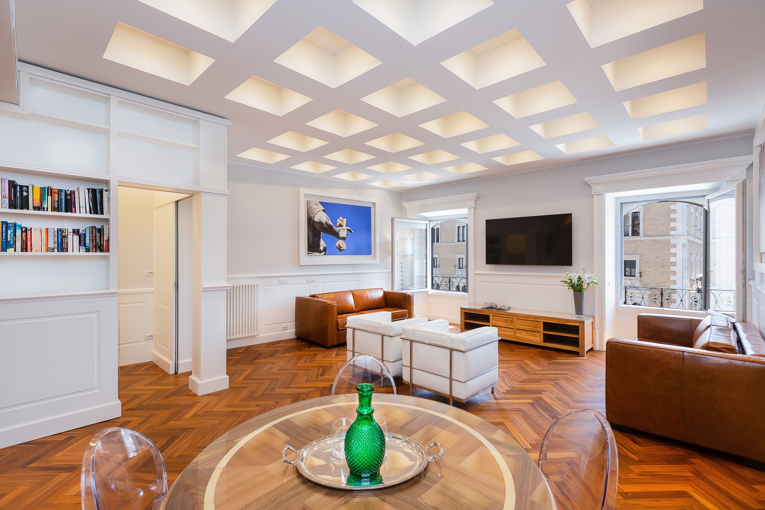A spacious living room with a wooden herringbone floor, white walls, and a ceiling with recessed lighting. There is a large flat-screen TV mounted on the wall, a brown leather sofa, and a white coffee table with a green vase.