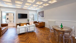 A spacious living room with a wooden herringbone floor, a large TV mounted on the wall, a brown leather sofa, a white sofa, and a dining table with clear chairs. - Amazing Vittoriano (Rome)