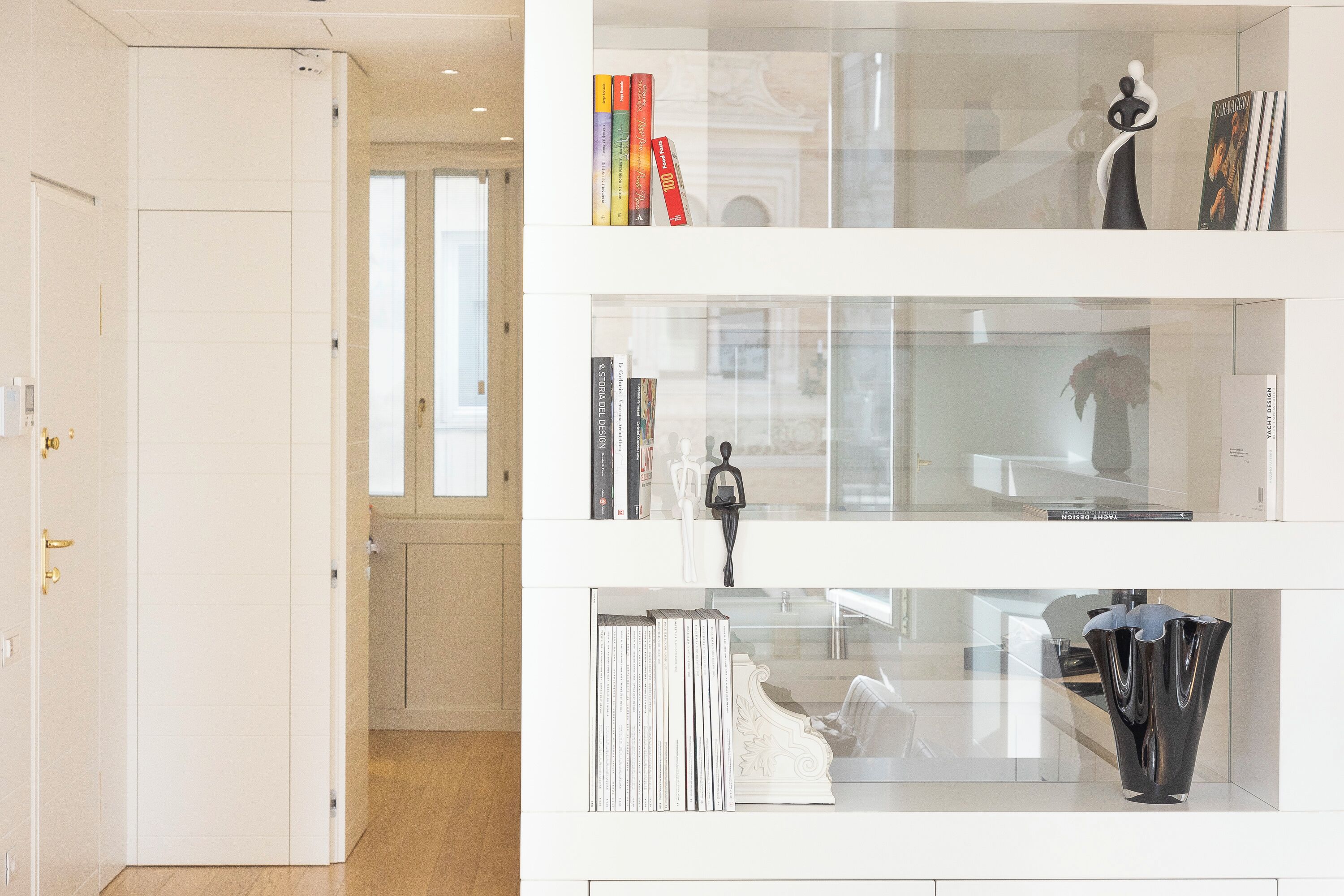 Un interior moderno con una puerta blanca, un pasillo y una vitrina de vidrio con libros y objetos decorativos.