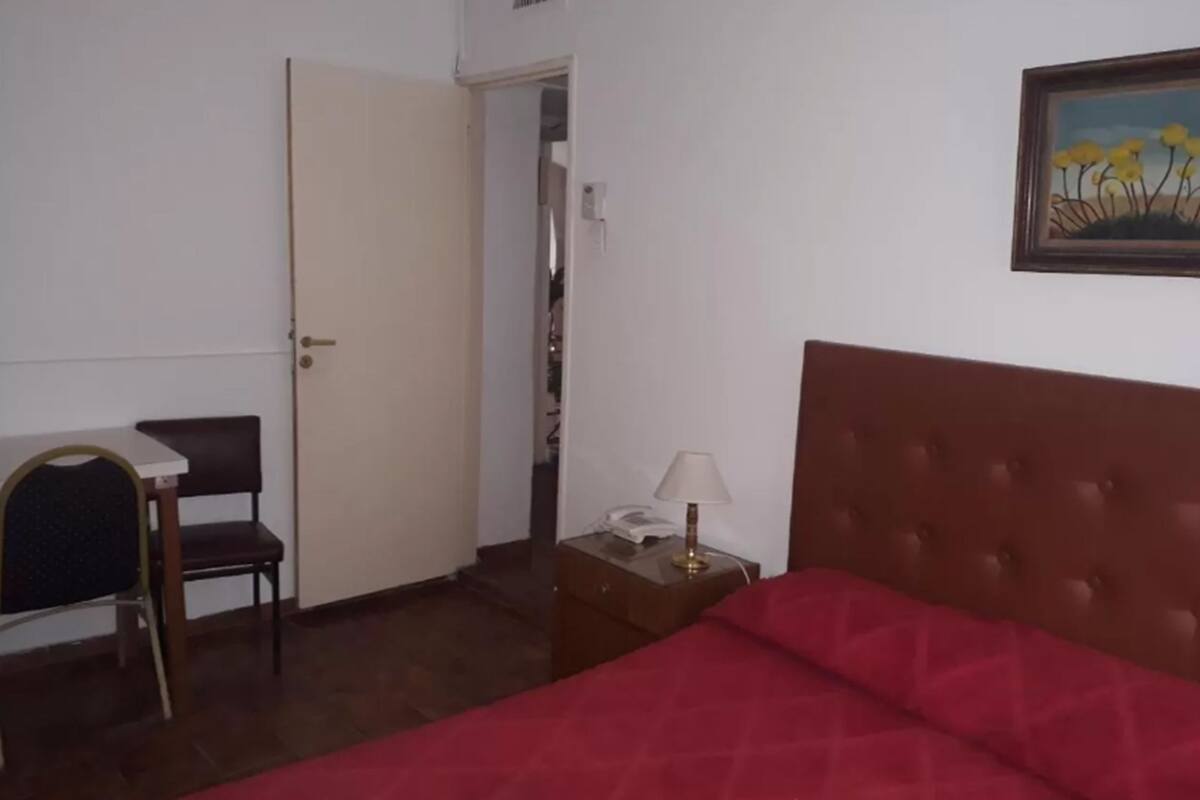A bedroom with a bed, a desk with a chair, a nightstand with a lamp, and a door leading to another room.