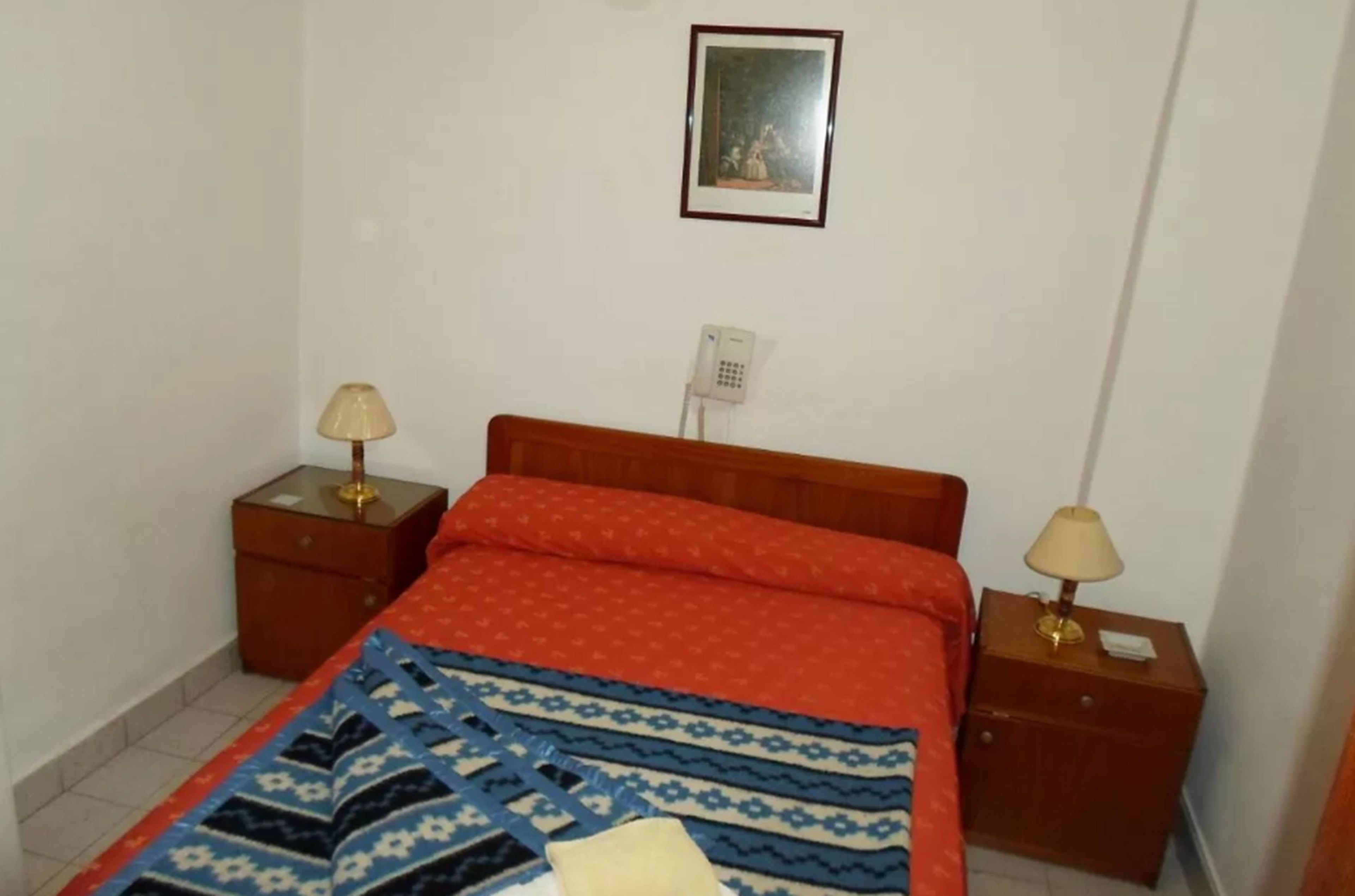 A bedroom with a wooden bed, two bedside tables, a lamp on each, a patterned bedspread, and a framed picture on the wall.