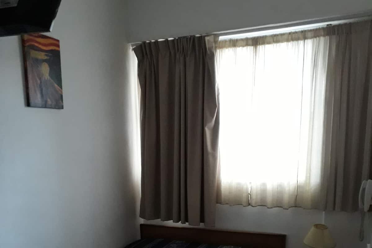 A bedroom with a bed, a nightstand, a lamp, a window with curtains, and a painting on the wall.