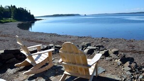 Two wooden rocking chairs on a rocky beach with a calm body of water and a forested island in the distance. - Sea-Breeze Cottage perched on the edge of the bay. (Eastport)