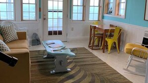 A room with a large window, a sofa, a dining table with chairs, and a small kitchen area. - Sea-Breeze Cottage perched on the edge of the bay. (Eastport)