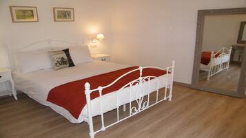 Un lit bien fait, avec une structure en métal blanc, une couverture rouge et deux oreillers. Il y a une table de chevet avec une lampe, et un grand miroir accroché au mur.