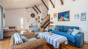 Een gezellige woonkamer met een blauwe bank, rieten meubels, een gestreept tafeltje en een houten trap.