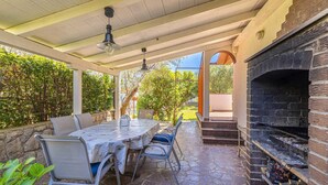 Een terras buiten met een stenen vloer, een tafel met stoelen, een barbecue en een pergola met hangende lampen.