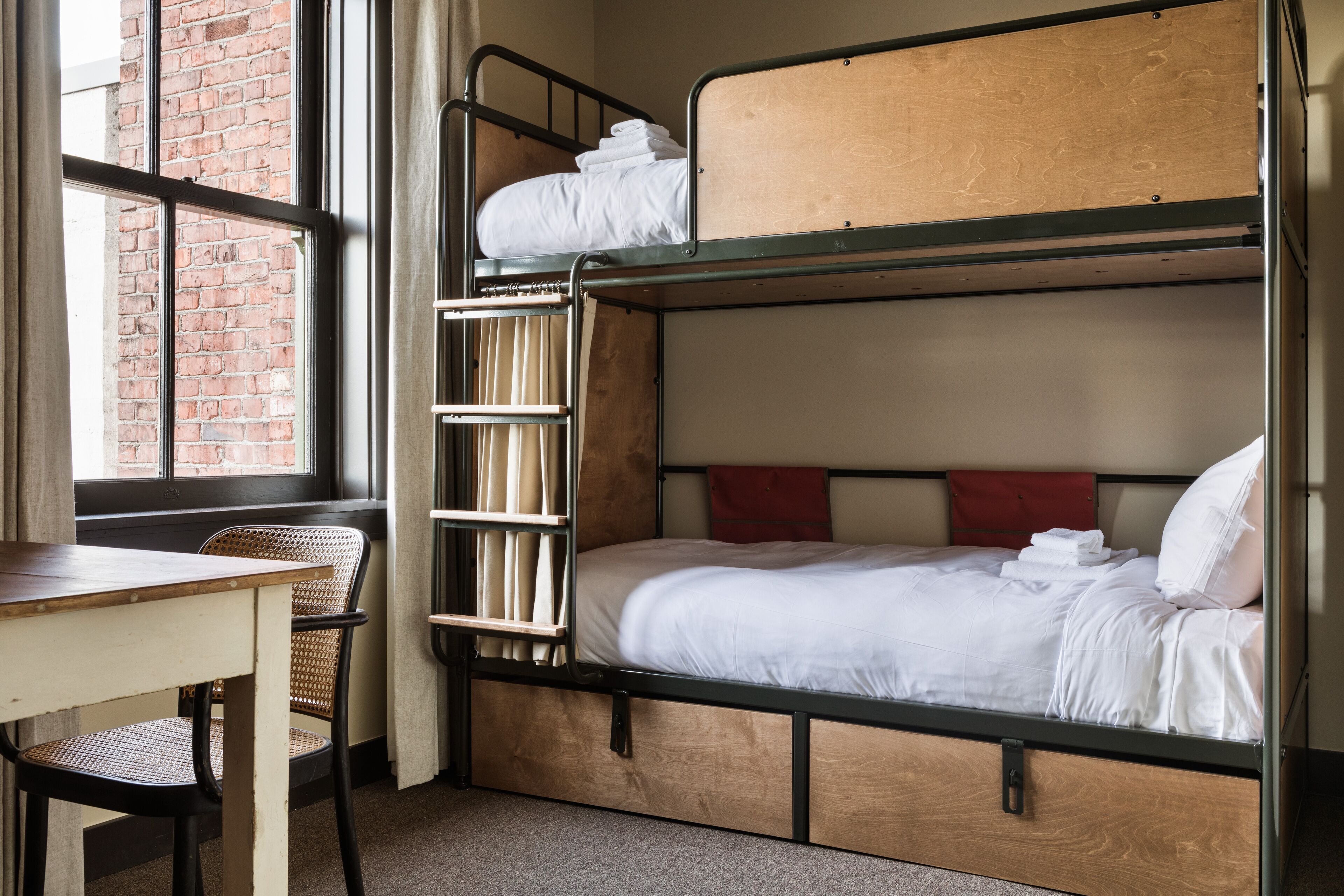 A bunk bed with a wooden headboard, a desk, and a chair in a room with a brick wall and a window.
