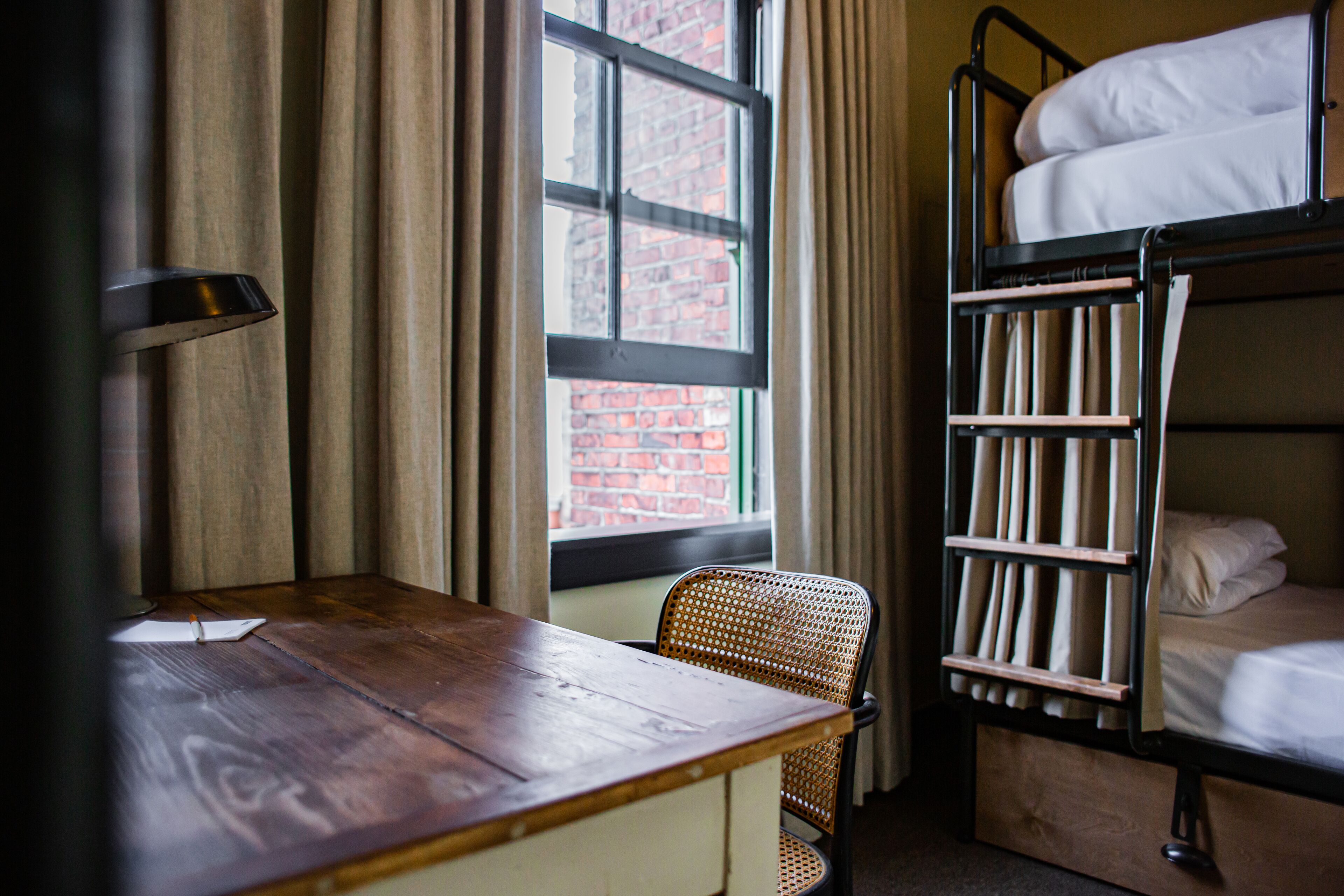 A bunk bed with a wooden ladder, a single chair, a wooden table, and a window with curtains.