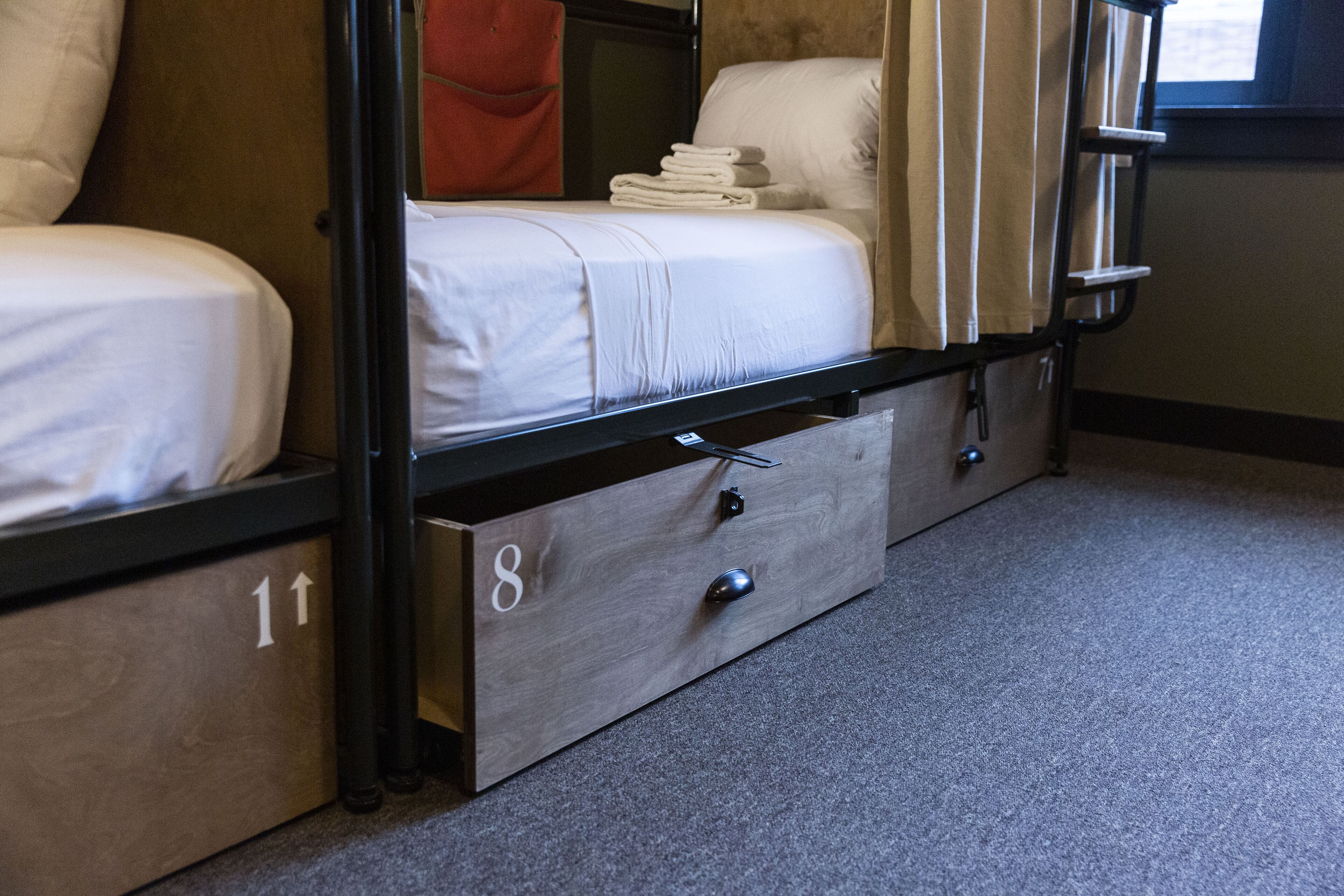 Una cama litera con estructura de madera, un colchón blanco y una toalla doblada con cuidado en la cama superior. Hay un cajón de madera pequeño con un pomo y el número "8" en el lateral.