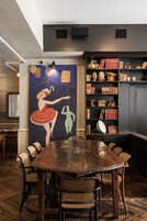 Un comedor con una mesa de madera y sillas de mimbre, una estantería con libros y objetos decorativos, y un mural en la pared.