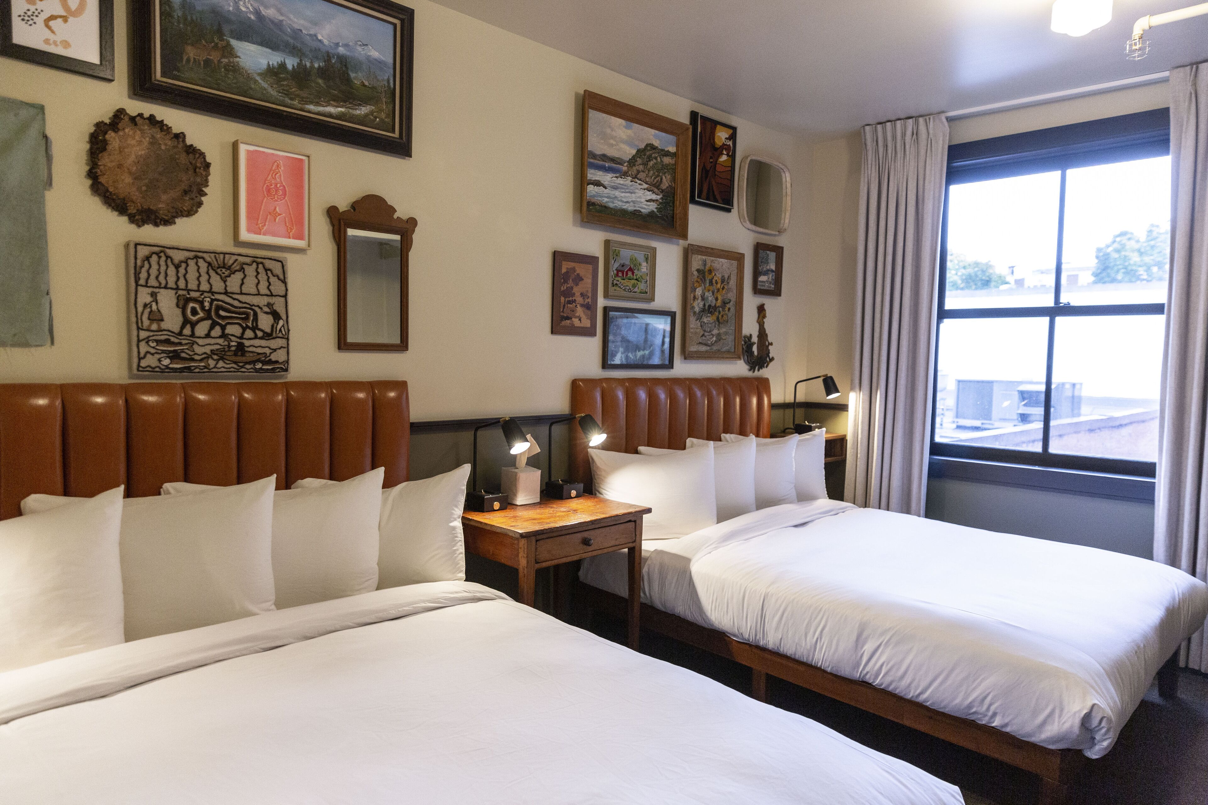 Habitación de hotel con dos camas, cabecero de madera, mesita de noche con lámpara y un ventanal con cortinas.
