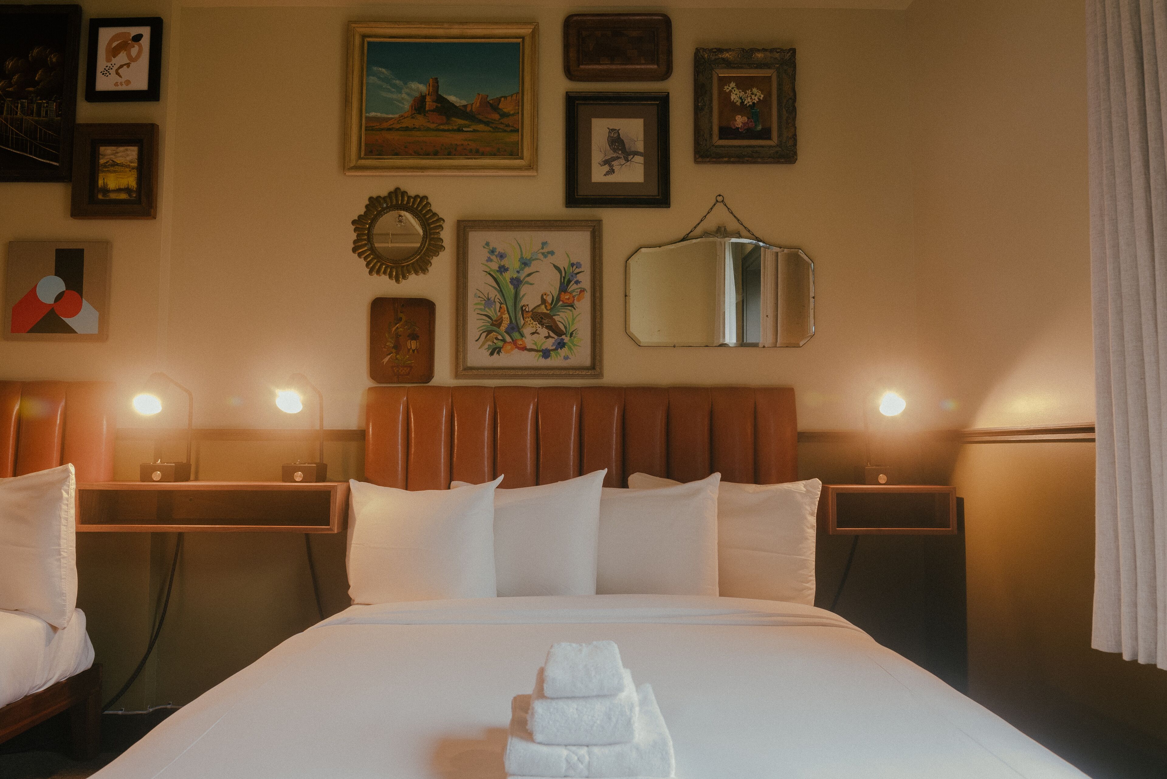 A hotel room with a neatly made bed, a bedside table with a lamp, and a wall decorated with framed pictures and a mirror.