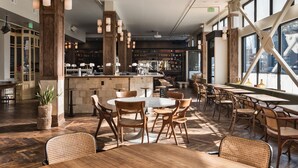 A spacious restaurant with wooden flooring, round tables, and wicker chairs. A bar area is visible in the background with stools.