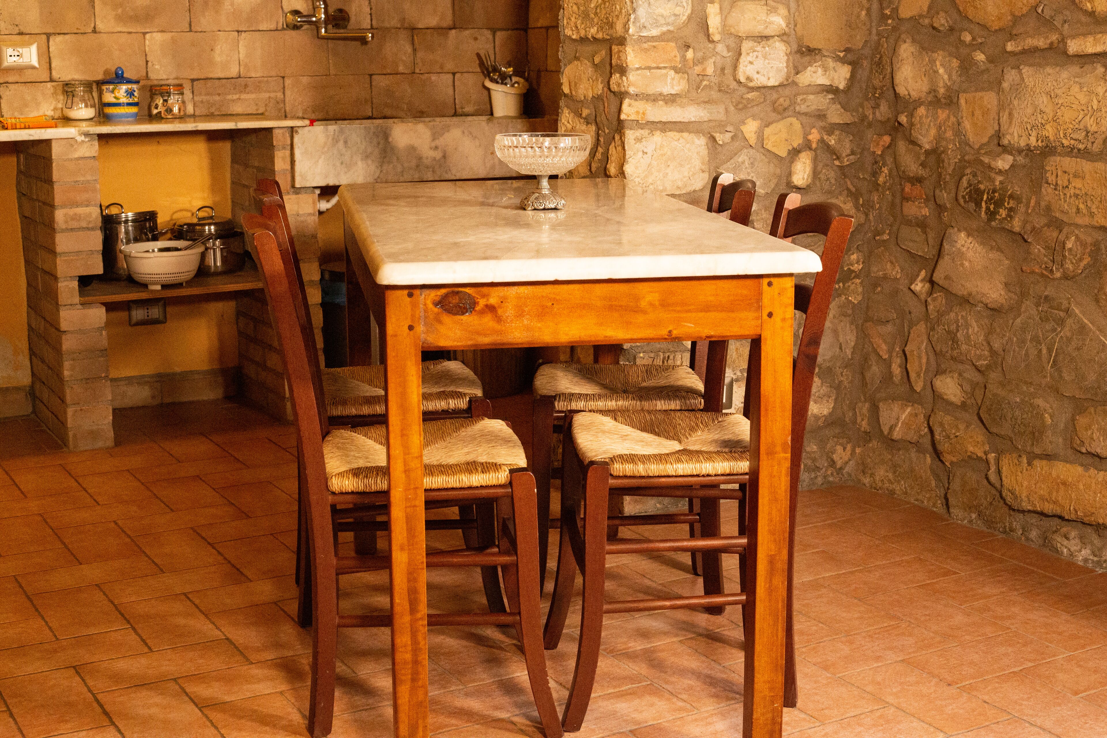 A wooden dining table with six chairs, a stone wall, and a kitchen area with shelves and utensils.