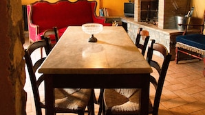 A cozy dining area with a wooden table, chairs, and a red sofa. A stone wall and a fireplace with a chimney are visible in the background. - Le Roghete (Acquapendente)