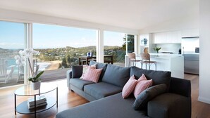 Una sala de estar moderna con un sofá gris, dos sillas blancas, una mesita redonda con una planta y vistas a una playa y colinas a través de amplios ventanales.