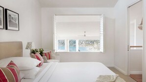 Un dormitorio con una cama grande, una ventana con persianas, una mesita de noche con lámpara y vistas a un paisaje costero.