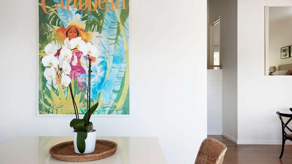 Une pièce avec un mur blanc, une œuvre d’art tropical encadrée, une plante en pot, une chaise en osier et une table à manger.